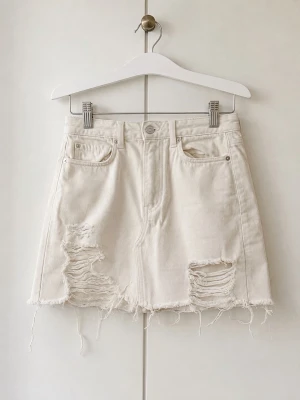Vit jeanskjol med slitningar - Säljer en vit kort jeanskjol med slitna detaljer och fransig nederkant. Kjolen har klassisk femficksmodell och råa kanter för en avslappnad look. Perfekt för dig som gillar en edgy och trendig stil.