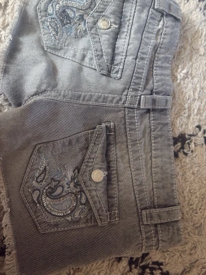 Grå jeansshorts med broderi - Säljer ett par grå jeansshorts med råa kanter och snygga broderade detaljer på bakfickorna. Shortsen har ficklock med knapp och bälteshällor. Perfekta för en avslappnad och trendig sommarlook.