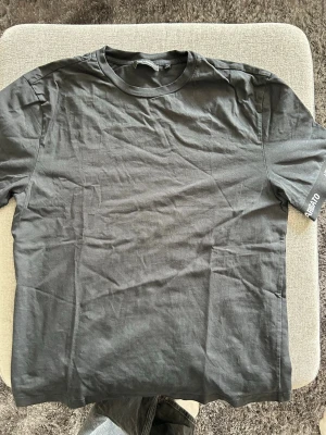 Svart t-shirt med Arigato-tryck - Snygg svart t-shirt med kort ärm och texten 'ARIGATO' broderad i vitt på ärmsluten. Klassisk rund halsringning och rak passform. Perfekt till jeans eller shorts för en clean och stilren look.