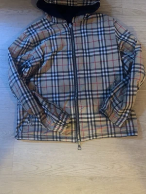 Reversibel burberry vindjacka  - Snygg beige vindjacka med klassiskt rutigt mönster i svart, vitt och rött. Jackan har dragkedja framtill, två fickor med dragkedja och en svart huva. Perfekt för dig som vill ha en stilren och trendig look.