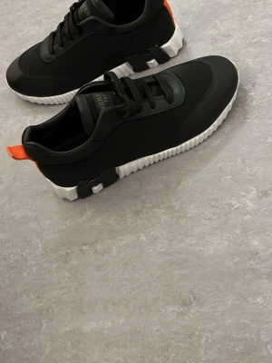 Svarta sneakers med vit sula - Snygga svarta sneakers med chunky vit sula och svarta detaljer undertill. Skorna har svarta snören och en orange flärp bak för enkel påtagning. Tillverkade i Italien och har en modern, sportig look med coola kontraster.