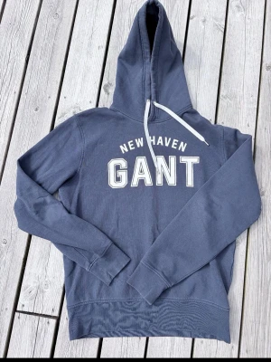 Blå hoodie från GANT med tryck - Snygg blå hoodie från GANT med vit text 'NEW HAVEN GANT' på bröstet. Klassisk huva med vit dragsko och långa ärmar. Perfekt för en avslappnad och trendig stil. Mjuk bomullskänsla och ribbade muddar vid ärmslut och nederkant.