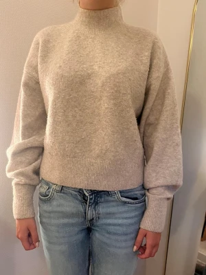 Beige stickad tröja med polokrage - Från En tjock mysig beige stickad tröja med hög polokrage och ballongärmar. Tröjan har breda ribbade muddar vid ärmslut och nederkant, vilket ger en avslappnad men ändå stilren look. Perfekt för kyliga dagar och enkel att matcha med jeans.