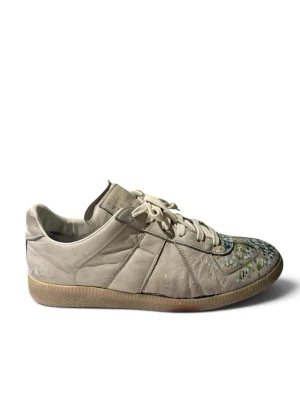 Maison Margiela sneakers med färgstänk - Beiga sneakers från Maison Margiela i läder med unika färgstänk i blått, grönt och vitt på tån. Klassisk låg modell med beige snörning och gummisula. Perfekt för dig som vill sticka ut med en kreativ och trendig look.storlek 40 1/2 passar 41