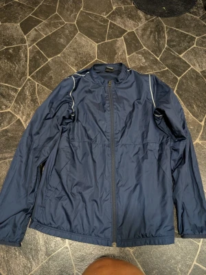 Mörkblå vindjacka från Nike XL. Barn 158-170 - Snygg mörkblå vindjacka från Nike i storlek XL. Jackan har hel dragkedja framtill, diskreta vita detaljer på axlarna och meshfoder på insidan för extra komfort. Perfekt för blåsiga dagar och sportiga outfits.
