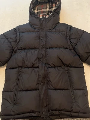 Svart pufferjacka från Burberry - Svart pufferjacka från Burberry med huva och dragkedja framtill. Jackan har två framfickor med dragkedja och ett stort Burberry London England-märke på ärmen. Fodret i huvan har rutigt mönster i grått, rött och vitt. Perfekt för kalla vinterdagar.