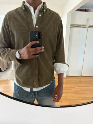 Olivgrön manchester overshirt - Säljer en olivgrön overshirt i manchester med klassisk krage och knappar framtill. Skjortan har lång ärm och en avslappnad passform, perfekt att bära över en t-shirt eller skjorta. Snygg och enkel att styla till jeans eller chinos.