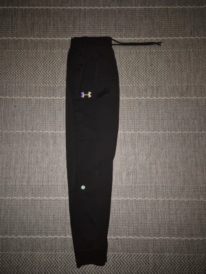 Under Armour Reflective - Svarta mjukisbyxor från Under Armour med resår och dragsko i midjan. Byxorna har muddar vid bensluten och en liten färgglad logga på ena benet. Perfekta för chill eller träning, tillverkade i mjukt material.