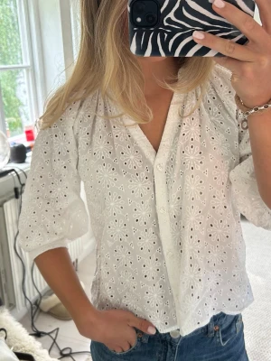 Vit broderad blus med trekvartsärm - Söt vit blus i broderad bomull med blommigt hålmönster och knäppning framtill. Blusen har trekvartsärmar med lätt puff och avslappnad passform. Perfekt till jeans för en fräsch och somrig look.