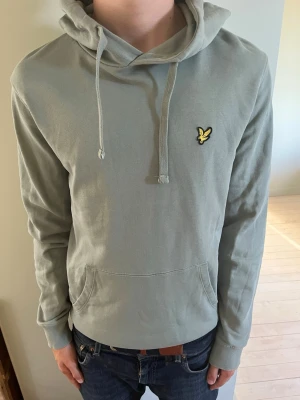 Grön hoodie med broderad logga - Snygg ljusgrå hoodie med huva och dragsnören. På bröstet sitter en broderad gul logga i form av en fågel. Hoodien har en klassisk känguruficka framtill och ribbade muddar vid ärmar och midja. Perfekt för en avslappnad och stilren look.          Skickas inom 1-3 dagar vid beställning 