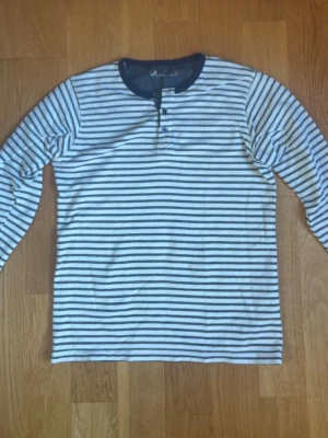 Randig långärmad tröja med knappar - Snygg långärmad tröja med smala marinblå och vita ränder från dressman. Tröjan har rund halsringning med tre knappar framtill och marinblå muddar vid ärmslut och hals. Perfekt för en avslappnad och stilren look. Storlek S