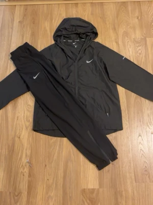 Svart Nike Running träningsset - Säljer ett svart träningsset från Nike Running med jacka och byxor. Jackan har huva, dragkedja och två fickor samt Nike-logga på bröstet. Byxorna har resår i midjan och vid benslut samt liten Nike-logga. Perfekt för löpning eller träning utomhus.