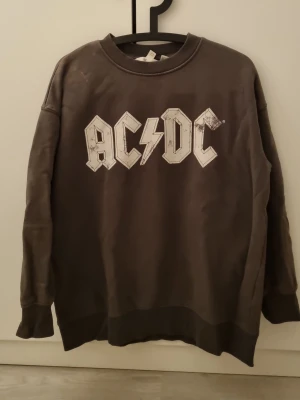 AC/DC sweatshirt - Säljer en mörkgrå sweatshirt från H&M med stort AC/DC-tryck i vitt på bröstet. Tröjan har rund halsringning, ribbade muddar vid ärmslut och nederkant samt en avslappnad passform. Perfekt för dig som gillar bandtröjor och vill ha en cool, casual stil.
