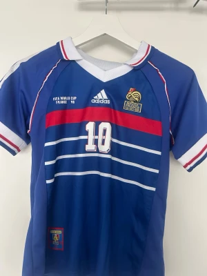 Fotbollströja + shorts Zidane - Klassisk blå fotbollströja och shorts från Spanien med Frankrikes landslagsmärke och nummer 10, Zidane, på ryggen. Tröjan har vita och röda detaljer, randigt mönster framtill och korta ärmar, aldrig använd