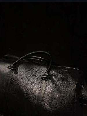 Classic black duffel bag ”HELT NY” - Stilren svart weekendbag i läderimitation med två handtag och justerbar axelrem. Väskan har en mjuk, rektangulär form och diskreta detaljer. Perfekt för dig som vill ha en enkel men snygg väska med plats för allt du behöver och kan användas till var som helst. Till exempel för att resa eller gå till gymmet.