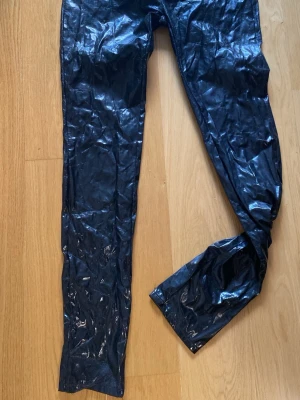 Svarta lackbyxor med snörning - Säljer ett par svarta leggings i glansigt material med snygg snörning framtill i midjan. De har en lös passform och ger en rockig look. Perfekta för dig som vill sticka ut lite extra på en spelning