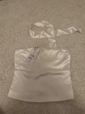 Vit satin bandeau topp med knytband - Säljer en snygg vit bandeau topp i satin med breda knytband som kan stylas runt halsen eller axlarna. Toppen har en glansig finish och är perfekt för dig som vill ha en trendig och elegant look.