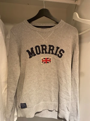 Grå tröja från Morris - Snygg grå tröja från Morris med mörkblå text och brittisk flagga på bröstet. Tröjan har rund halsringning, ribbade muddar och är tillverkad i mjukt stickat material. Perfekt för en avslappnad och stilren look.