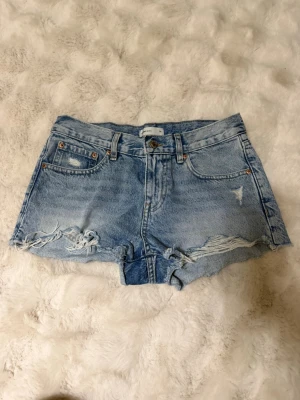 Blå jeansshorts med slitningar - Säljer ett par ljusblå jeansshorts från perfect jeans i storlek 34. Shortsen har råa kanter, slitna detaljer och klassisk femficksmodell. Perfekta för sommaren och ger en avslappnad vibe till din outfit.