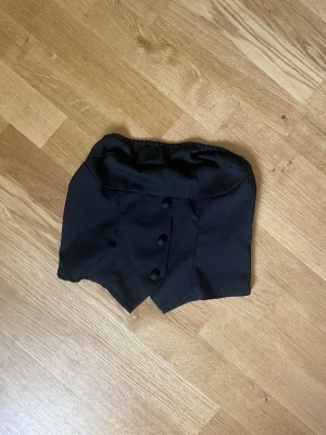 Svart korsett-tubtopp med knappar - Snygg svart korsett-inspirerad tubtopp med dekorativa knappar framtill och dragkedja bak. Toppen har en unik spetsig nederkant och elastisk kant upptill för bra passform. Perfekt för att skapa en edgy och trendig look.