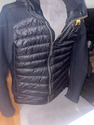 Svart hybrid pufferjacka med dragkedja - Snygg svart pufferjacka med quiltad front och rygg i glansigt material samt stickade ärmar och sidor. Jackan har hel dragkedja framtill, hög krage och en praktisk ficka med dragkedja bak. Perfekt för kalla vinterdagar och ger en sportig look.