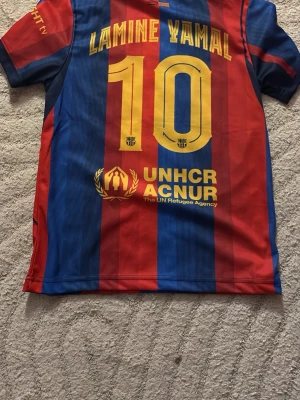 Barcelona matchtröja Lamine Yamal #10 från utomlands  - Säljer en officiell FC Barcelona matchtröja med Lamine Yamal och nummer 10 på ryggen. Tröjan har klassiska blå och röda vertikala ränder, gula detaljer och klubbmärke på bröstet. Tillverkad i lätt och ventilerande material, perfekt för fotbollsträning eller match. Sätter är ifrån utomlands och shortsen är jätte stretchiga så är mycket större i midjan än vad de ser ut 