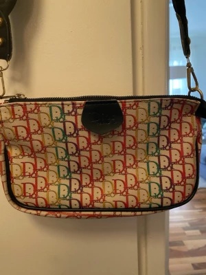 Flerfärgad monogram handväska - Snygg handväska med färgglatt monogrammönster i rött, grönt, gult och lila på vit bakgrund. Väskan har svarta detaljer och axelrem i svart material. Perfekt för dig som vill sticka ut med en unik accessoar.
