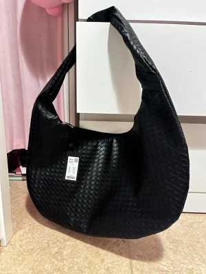 Svart axelväska med flätad struktur - Helt ny snygg svart axelväska i konstläder med flätad struktur och bred axelrem. Väskan har en mjuk, rundad form och stängs med dragkedja. Perfekt för dig som vill ha en trendig och rymlig väska till alla dina grejer.