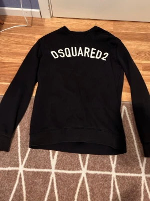 Svart sweatshirt från Dsquared2 - Cool svart sweatshirt från Dsquared2 med vit logga tryckt över ryggen. Klassisk rund halsringning och långa ärmar. Tillverkad i mjukt material som känns skönt mot huden. Perfekt för en avslappnad och trendig look.
