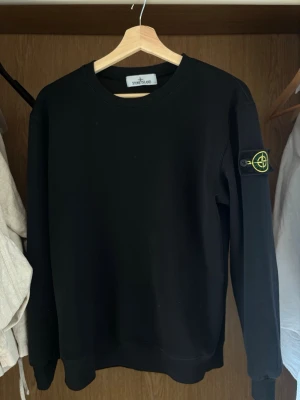 Svart sweatshirt från Stone Island - Säljer en svart sweatshirt från Stone Island med den klassiska loggan på vänster ärm. Tröjan har rund halsringning och ribbade muddar vid ärmslut och nederkant. Perfekt för dig som gillar stilrena och ikoniska plagg. Pris kan diskuteras 