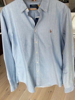 Ljusblå skjorta från Polo Ralph Lauren - Klassisk ljusblå skjorta från Polo Ralph Lauren med broderad logga på bröstet. Skjortan har knappar framtill, lång ärm. Storleken är L men kan passa stor M också. Köpt i USA (newyork) ny i RL butik men var för stor.  Köpte den för 130 $ så rejäl prissänkning. 