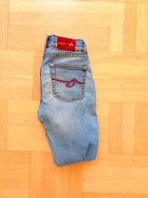 Ljusblå jeans från Jacob Cohen - Snygga ljusblå jeans från Jacob Cohen med röd broderad detalj på bakfickan och patch i rött på linningen. Tillverkade av japanskt denimtyg och har klassiska kontrastsömmar. Perfekta för dig som gillar en unik touch på dina jeans. Priset inte hygget i sten ett par riktigt fina jeans om de passar dig tveka inte på att höra av dig vid minsta fråga! 😄😁Nypriss 5500kr 