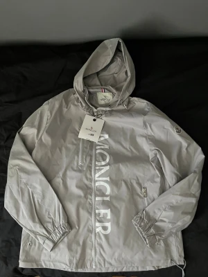 Moncler Grå Vindjacka - Snygg grå vindjacka från Moncler med stor logga framtill. Jackan har huva, dragkedja, två sidofickor med dragkedja och elastiska muddar vid ärmsluten. Tillverkad i lätt och tåligt material, perfekt för blåsiga dagar. Klassisk Moncler-patch på ärmen.     (PRIS KAN DISKUTERAS)