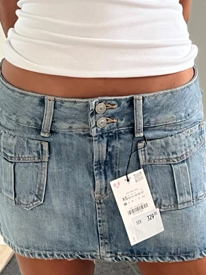 Blå jeanskjol med framfickor - Säljer en klassisk blå jeanskjol i kort modell med två knappar i midjan och framfickor samt designade slitningar. Kjolen har bälteshällor och en lågmidjad passform. Perfekt till vardags för en avslappnad och trendig look.