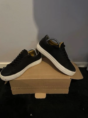 Svarta låga sneakers med vit sula - Stilrena svarta sneakers med lågt skaft och vit, tjock sula. Skorna har svarta skosnören och en enkel, klassisk design. Insidan har en kontrasterande gul detalj. Passar perfekt till streetwear och vardagsoutfits.