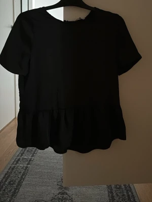 Svart blus med volangkant - En svart blus med korta ärmar och rund hals. Blusen har en volangkant nedtill som ger en söt och trendig look. Perfekt att matcha med jeans eller kjol för en enkel men snygg outfit.