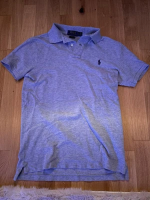 Grå pikétröja från Polo Ralph Lauren - Klassisk grå pikétröja från Polo Ralph Lauren i slim fit-modell. Tröjan har korta ärmar, krage och knappar framtill samt den ikoniska broderade loggan på bröstet. Tillverkad i mjuk bomull för en bekväm känsla.
