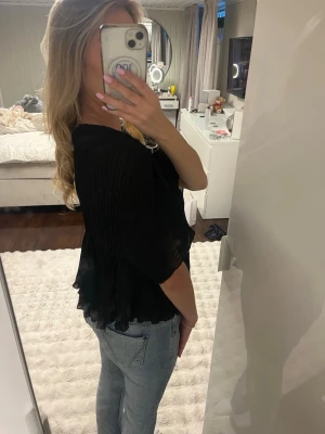 Svart blus med volang och v-ringning - Säljer en svart blus med v-ringad hals och volangdetaljer nedtill. Blusen har korta, lösa ärmar och ett lätt, luftigt tyg som ger en flowy känsla. Perfekt att styla med jeans eller kjol för en trendig look.