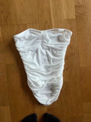 Vit axelbandslös topp med mesh - Säljer en vit axelbandslös topp med rynkad mesh över hela toppen. toppen har en tight passform och är lätt transparent för en trendig look. Perfekt för dig som vill ha en enkel men snygg outfit.