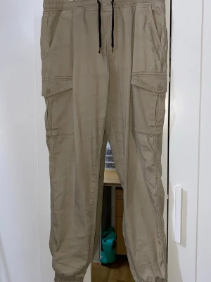Beige cargobyxor med dragsko - Snygga beige cargobyxor med elastisk midja och svart dragsko. Byxorna har stora sidofickor med lock och knapp, samt muddar vid bensluten. Perfekta för en avslappnad och trendig stil. Tillverkade i ett mjukt bomullsliknande material.