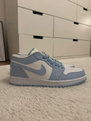 Nike Air Jordan 1 Low blå/vit - Snygga Nike Air Jordan 1 Low sneakers i ljusblått och vitt. Skorna har klassisk siluett med perforerad tåbox, blå swoosh och matchande blå snörning. Tillverkade i läder med textildetaljer och ikoniska Jordan-loggor på plös och häl. Perfekta för dig som gillar streetwear. Helt nya aldrig använda. 