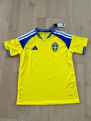 Svensk landslagströja Adidas gul/blå - Gul svensk landslagströja från Adidas med blå detaljer och vita ränder på axlarna. Tröjan har korta ärmar, broderad landslagssköld på bröstet och Adidas-logga. Tillverkad i lätt och funktionellt material, perfekt för fotboll eller träning.