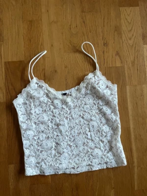 Vit spetslinnetopp med blommönster - Söt vit linnetopp med tunna axelband och blommigt spetsmönster. Toppen är ärmlös och har en lätt transparent look som ger en romantisk vibe. Perfekt att styla med jeans eller kjol för en trendig outfit.