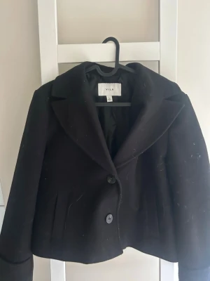 Svart kappa från VILA med stora slag - Stilren svart kappa från VILA med stora slag och två knappar framtill. Kortare modell med raka ärmar och diskreta fickor på sidorna. Perfekt för dig som gillar en klassisk look med modern twist.