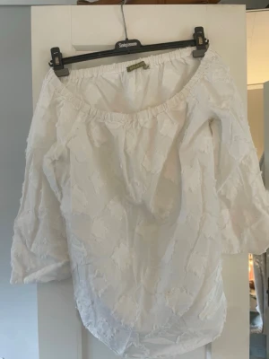 Vit offshoulder blus med struktur - Säljer en vit offshoulder blus med trekvartsärm och elastisk halsringning. Blusen har ett unikt strukturerat tyg med fransiga fyrkantiga detaljer som ger en cool och bohemisk känsla. Perfekt till jeans eller kjol för en avslappnad look.