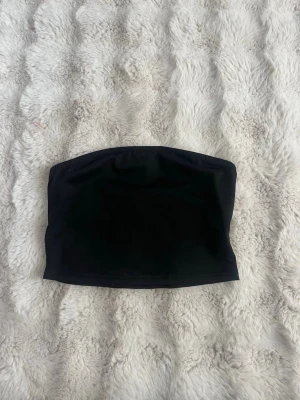 Svart bandeau tubtopp - Enkel och stilren svart bandeau tubtopp i stretchigt material. Toppen är axelbandslös och har en rak passform som sitter tight mot kroppen. Perfekt att matcha med höga jeans eller kjol för en trendig look.
