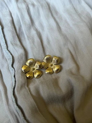 Guldiga blomformade örhängen - Säljer ett par örhängen i metall med blommig design och guldfärgad finish. Örhängena har fem kronblad med fina räfflade detaljer som ger en elegant och unik look. Perfekta för dig som vill ha något som sticker ut och känns trendigt.