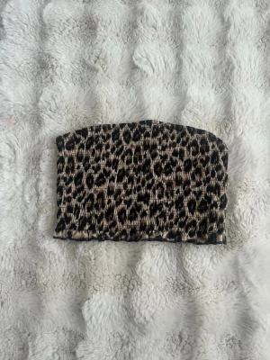 Leopardmönstrad bandeau tubtopp - Säljer en leopardmönstrad bandeau tubtopp i beige och svart. Toppen är smockad och har en elastisk passform som sitter snyggt på kroppen. Perfekt för dig som vill ha en trendig och djurinspirerad look.