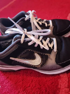 Svarta och grå Nike sneakers - Snygga sneakers från Nike i svart och grått med vita detaljer och klassisk swoosh-logga på sidan. Skorna har vita snören och en låg profil. Ovandelen är i mesh och syntetmaterial för bra andningsförmåga och komfort.