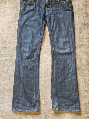 Mörkblå bootcut ola - Säljer ett par mörkblå jeans med raka ben och klassiska kontrastsömmar. Jeansen har fem fickor och är tillverkade i kraftigt denimtyg. Perfekta för dig som gillar en tidlös och enkel stil. Modellen på nudie jeansen är bootcut ola med storleken 30/32